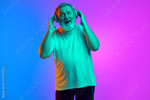 Emotional, bearded, senior man in casual clothes listening to music in headphones on gradient blue-purple background in neon light. Concept of human emotions, lifestyle, casual fashion