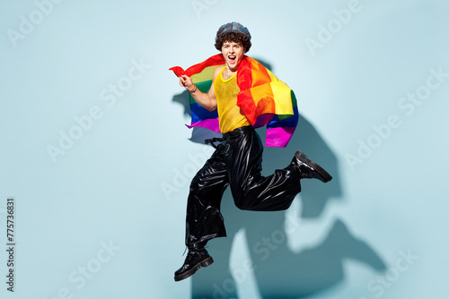 Full body side view young gay man wears mesh tank top hat clothes wrapped in striped rainbow flag jump high isolated on plain blue background studio portrait. Pride day June month love LGBT concept. photo