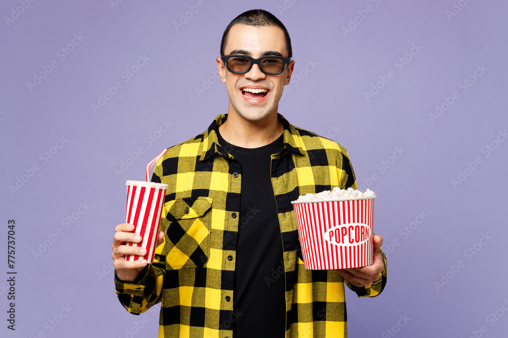 Fototapeta premium Young smiling fun middle eastern man wear yellow shirt casual clothes 3d glasses watch movie film hold bucket of popcorn cup of soda pop in cinema isolated on plain purple background studio portrait.