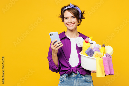 Young happy woman wear purple shirt hold basin with detergent bottles do housework tidy up hold in hand use mobile cell phone isolated on plain yellow background studio portrait. Housekeeping concept.
