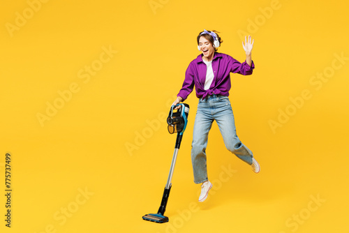 Full body young woman wear purple shirt casual clothes do housework tidy up hold vacuum cleaner listen music in headphones leap bounce isolated on plain yellow background studio. Housekeeping concept. photo