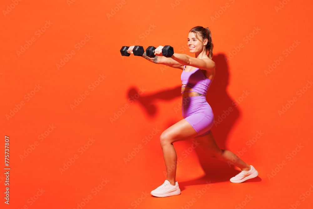 Full body young fitness instructor woman sportsman wear top shorts purple clothes train in home gym hold dumbbells do squats lunges isolated on plain orange background. Workout sport fit abs concept. Full body young fitness instructor woman sportsman wear top shorts purple clothes train in home gym hold dumbbells do squats lunges isolated on plain orange background. Workout sport fit abs concept.