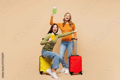 Young friends two women wearing orange green shirt together hold passport ticket sit on bag isolated on plain beige background. Tourist travel abroad in free time rest getaway Air flight trip concept