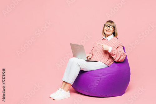 Full body elderly IT woman 50s year old wear sweater shirt casual clothes glasses sit in bag chair hold use work point finger on laptop pc computer isolated on plain pink background Lifestyle concept