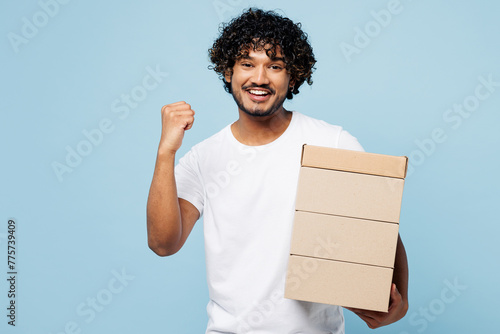 Young happy Indian man he wear white t-shirt casual clothes hold stack cardboard blank boxes do winner gesture isolated on plain pastel light blue cyan background studio portrait. Lifestyle concept.