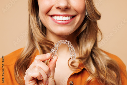 Close up cropped young happy woman wear orange shirt casual clothes hold in hand invisible transparent aligners, invisalign bracer isolated on plain pastel light beige background. Lifestyle concept. photo