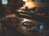 A dimly lit living room scene with a person relaxing on a couch, amidst scattered evidence of solitary indulgence in whiskey and cigarettes.

