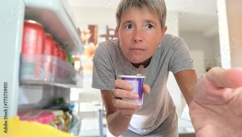 Seen from inside the refrigerator, an adult woman with short gray hair, with a determined and serious look,holding a jar of pudding with a teaspoon inside and taking something else with the other hand