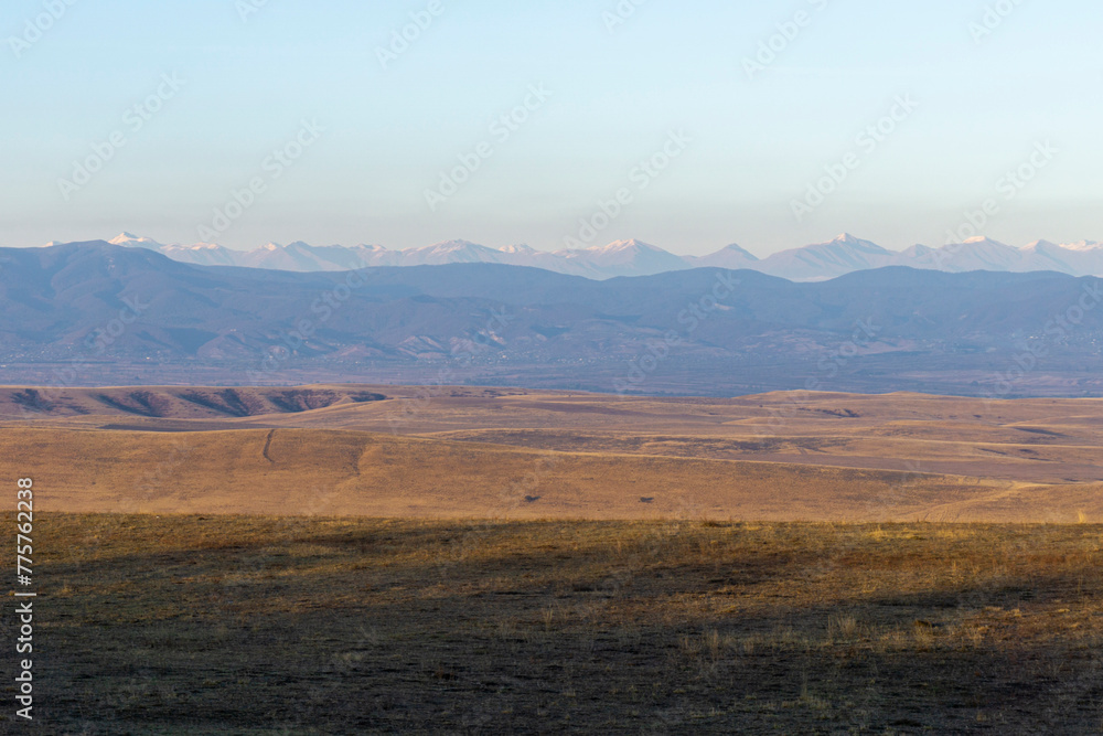 Landscape view of savanna mountains and snow-capped Caucasus ridge. Dried orange grass. Bright blue sky. Desert region in Georgia.