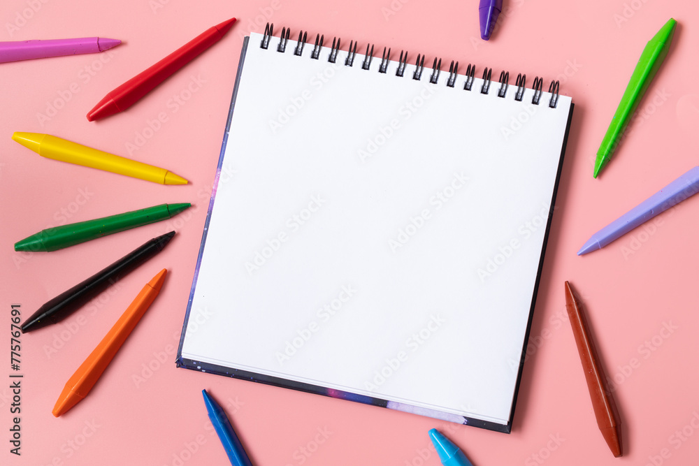 Open sketchbook with white empty blank. Colorful wax crayons on the pastel pink background. Flat lay composition. Open sketchbook with white empty blank. Colorful wax crayons on the pastel pink background. Flat lay composition.