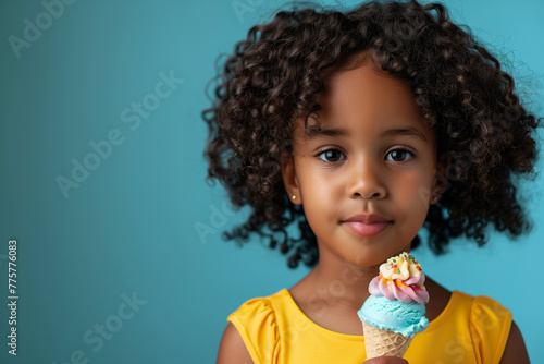 A young black girl in a yellow dress enjoys a colorful ice cream cone against a blue background