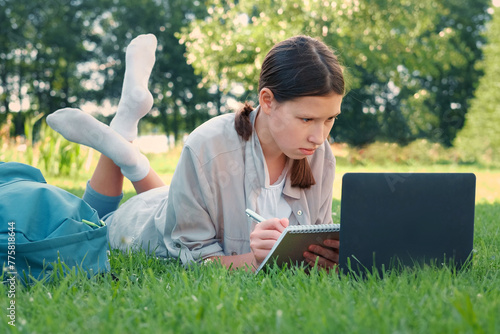 Teenage schoolgirl studying reading her books, tablet and notebook, sitting outdoors. Back to school. Student girl lying on the green grass using laptop in the college yard or park. Distance learning.