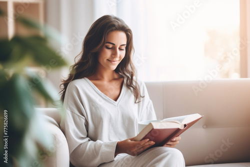 Pensive relaxed Latin woman reading a book at home, drinking coffee sitting on the couch. Copy space