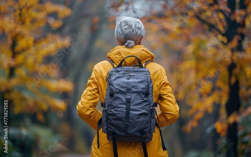 A woman wearing a yellow jacket and carrying a backpack is walking through a forest. The autumn leaves are falling around her, creating a serene and peaceful atmosphere photo
