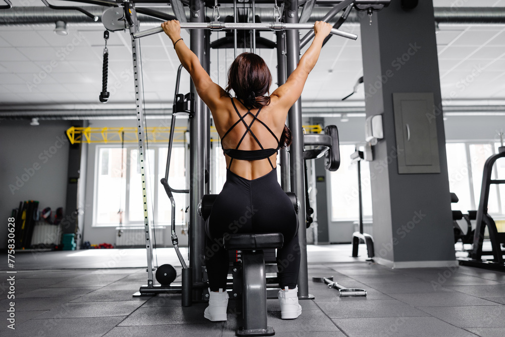 Fototapeta premium Muscular athletic female bodybuilder in black suit pulled on sports simulator in gym. Back muscles are very tense.