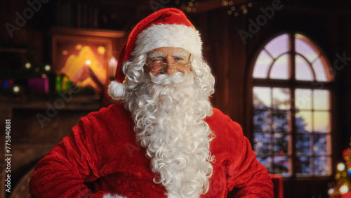 Portrait of Santa Claus Standing in a Festive Cozy Home with Fireplace, Decorations, Snow Falling Outside, Christmas Tree. Santa Looking at Camera, Smiling, Winking. Winter Holiday Celebrations