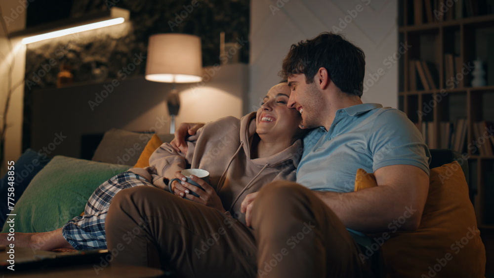 Smiling spouses watching tv sitting at cozy living room with coffee late evening