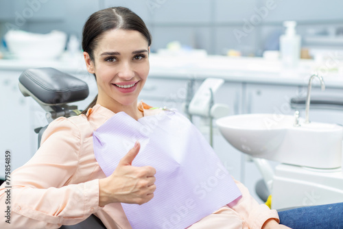 Satisfied woman patient showing her perfect smile and gesturing thumb up after treatment, sitting in dentist clinic photo