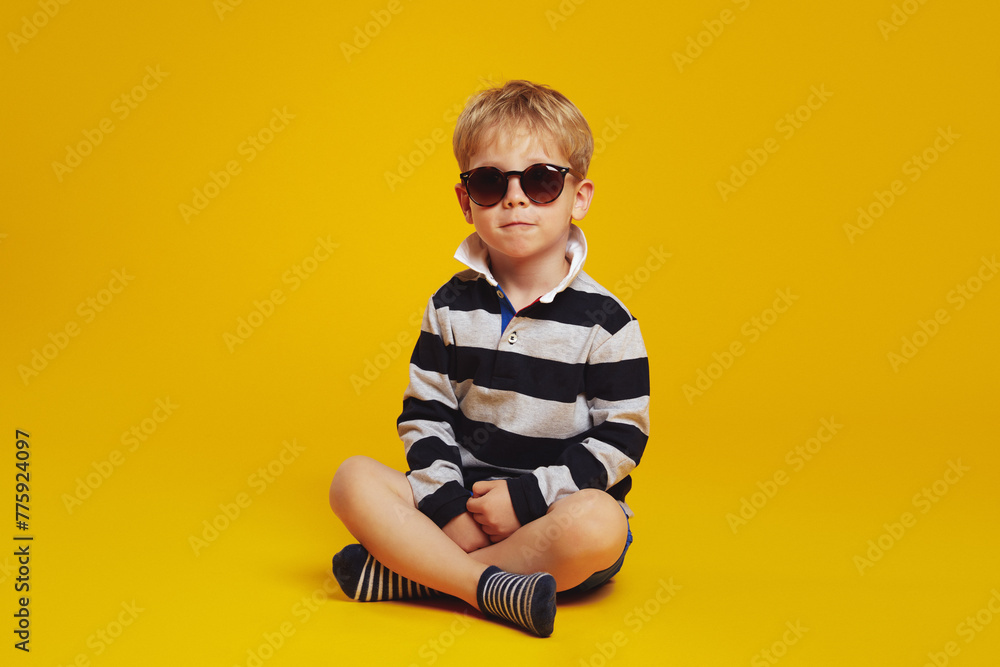 Fototapeta premium Cool little serious boy in striped shirt and trendy sunglasses feels confident while sitting crossed legged on floor against the yellow background