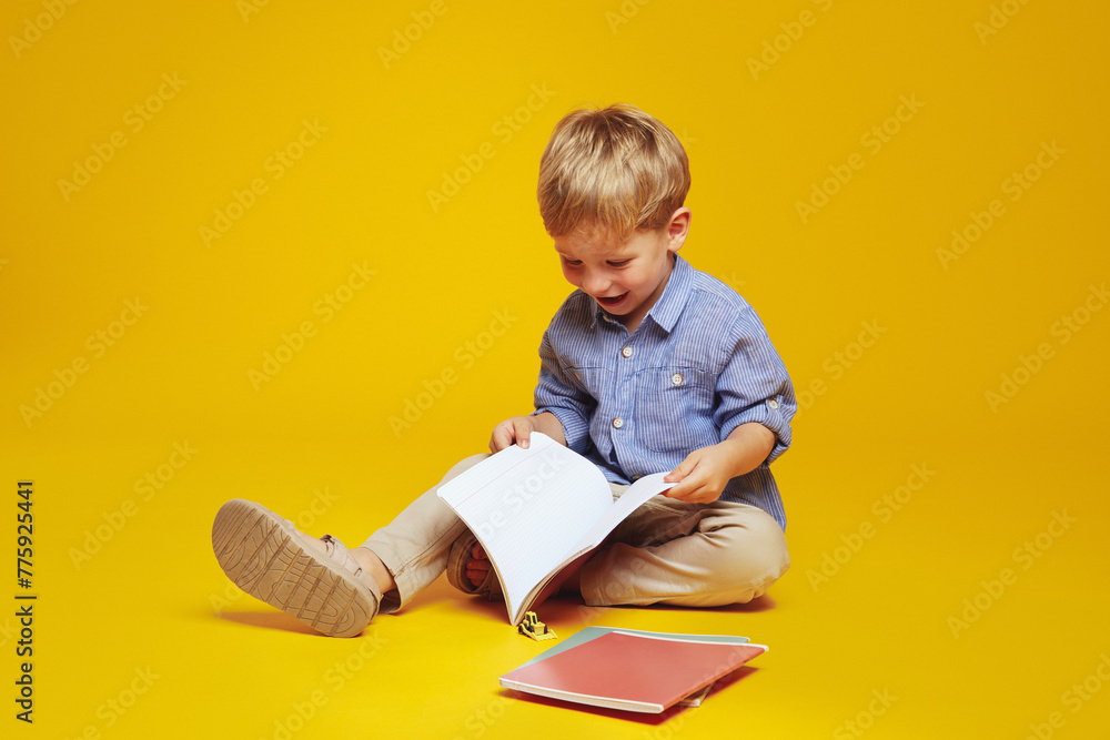 Positive curious schoolboy in casual clothes sitting on studio floor and reading notepad while smiling excitedly, isolated over yellow background. Positive curious schoolboy in casual clothes sitting on studio floor and reading notepad while smiling excitedly, isolated over yellow background.