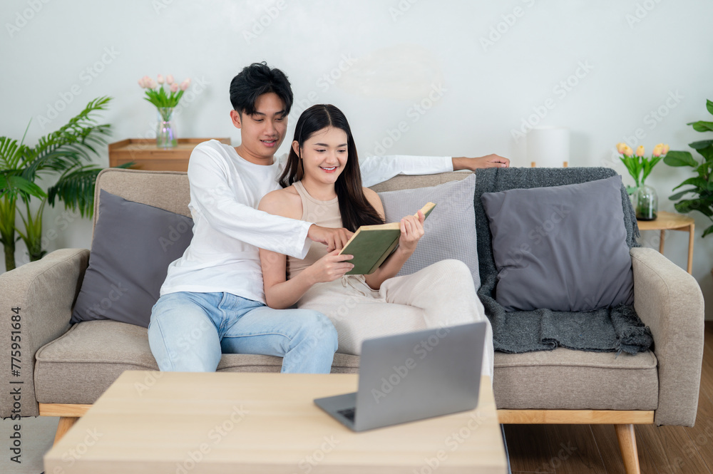 A happy Asian couple shares a moment reading a book together with a laptop, blend of traditional and digital pastimes.