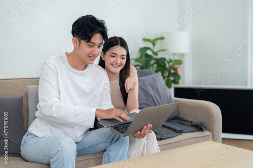 An Asian couple smiling and engaging with a laptop screen while sitting on a sofa in a cozy living room environment.