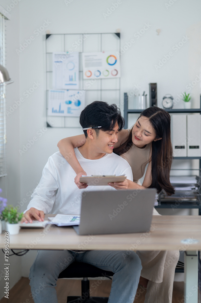 A smiling young Asian couple collaborates on a project, works together managing their budget, working on a laptop in a home office setting.