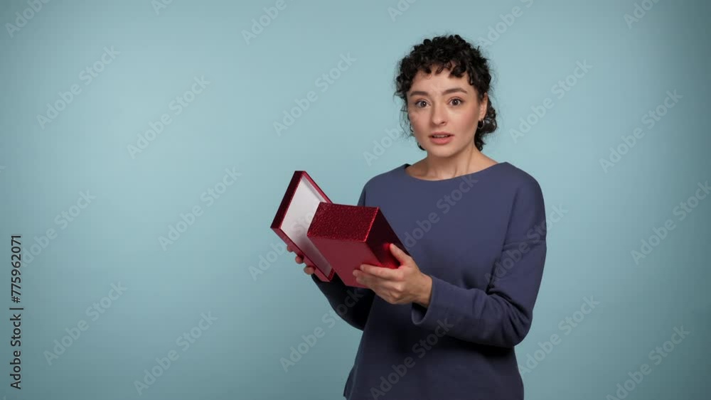 Shocked curly woman opens present red box surprised looks camera and says wow. Excited positive female wearing blue sweater with gift in hands on isolated light blue background with copy space Shocked curly woman opens present red box surprised looks camera and says wow. Excited positive female wearing blue sweater with gift in hands on isolated light blue background with copy space