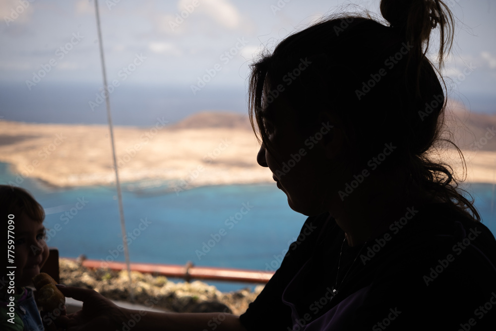 Mother and daughter silhouette against the light looking at the island of La Graciosa from the viewpoint of El Rio. Turquoise ocean. Blue sky with big white clouds. Lanzarote, Canary Islands, Spain Mother and daughter silhouette against the light looking at the island of La Graciosa from the viewpoint of El Rio. Turquoise ocean. Blue sky with big white clouds. Lanzarote, Canary Islands, Spain
