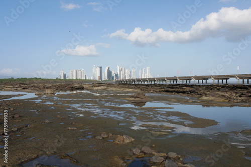 Wattenmeer an der Küste von Panama-Stadt bei Niedrigwasser am Meer mit Felsen und der als Hochstraße ausgeführten Umgehungsstraße Corredor Sur sowie die Skyline mit Hochhäusern im Hintergrund
