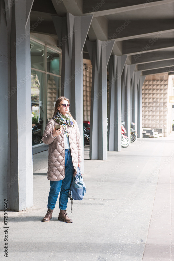 Beautiful stylish young woman with bag, sunglasses and mobile phone standing near building on the street