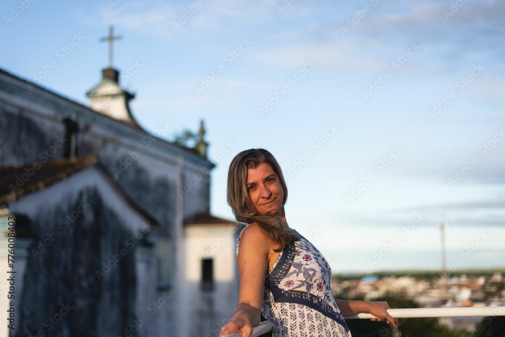 Fototapeta premium Woman on the porch of her house looking at the camera against the sky and church in the background