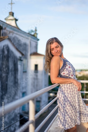 Woman on the porch of her house looking at the camera against the sky and church in the background