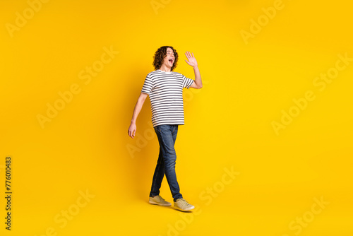 Full size portrait of nice young man walk empty space arm wave hi wear striped t-shirt isolated on yellow color background