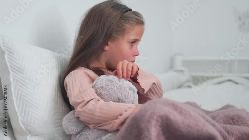 Upset sick girl coughing while sitting with soft toy on bed at home. Sad girl child suffering from cold and flu and using digital tablet in apartment. photo