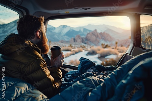 A man sitting in the trunk of his car, holding coffee and looking at mountains through an open window with blue blankets on him, camping gear inside, sunset light © Svetlana