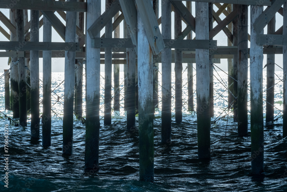 Capturing the intricate patterns of light and shadow, this image showcases the robust wooden structure of a pier with its pillars and crossbeams plunging into the dark, swirling waters below, offering