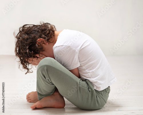A young boy is seated on the wooden floor with head bowed down