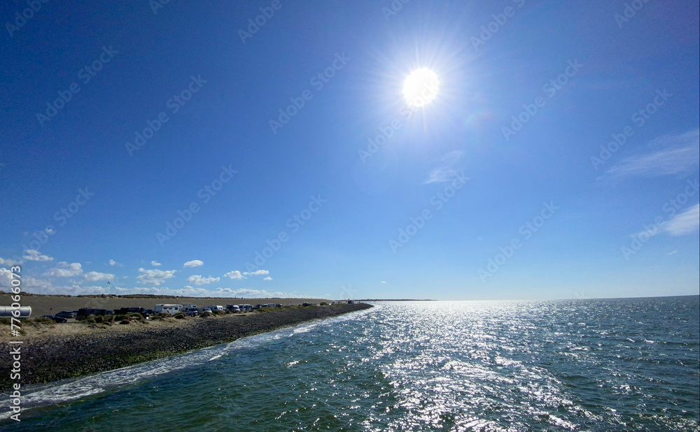 This vibrant image captures a bright sunny day at the seaside, featuring a glistening sea reflecting the sunlight, a clear blue sky, and a stony shoreline that leads to a distant horizon. The sun's