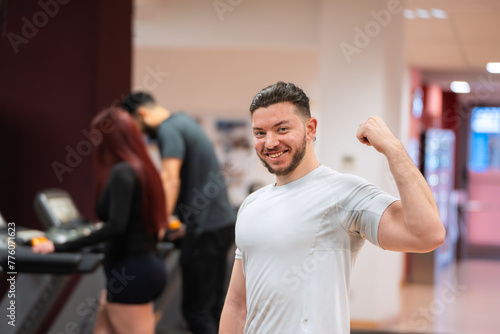 Muscular man in white tee flexes his bicep  with focused gym-goers behind him. 