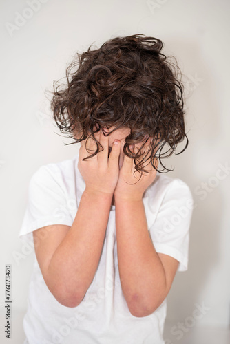 A young boy with curly black hair covering his face with his hands