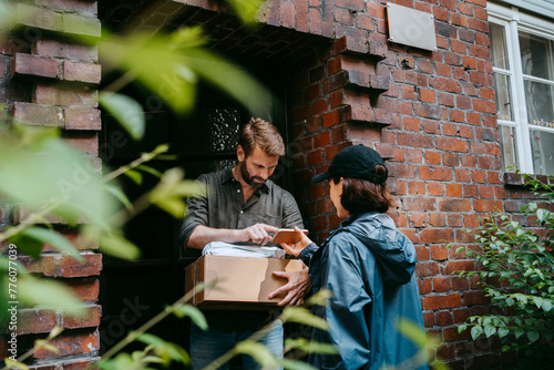 Mature man doing electronic signature while receiving parcel from female delivery person standing at doorstep photo