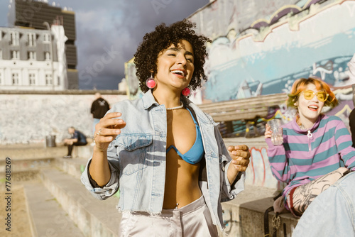 Carefree smiling non-binary person dancing with friends sitting on steps in city at sunny day photo