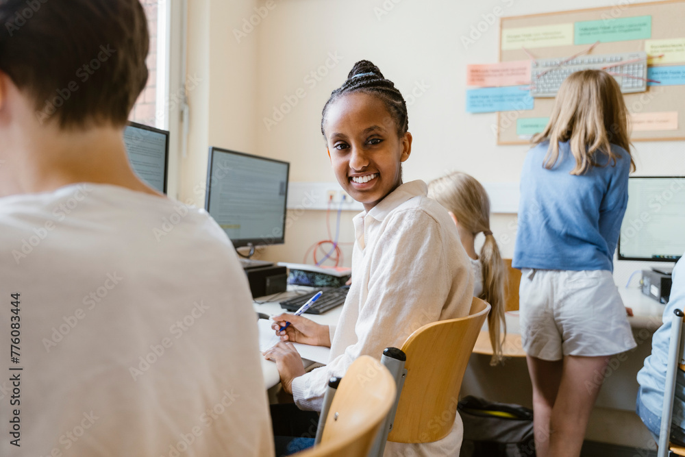 Side view portrait of smiling girl sitting on chair with friends in computer class at school Side view portrait of smiling girl sitting on chair with friends in computer class at school