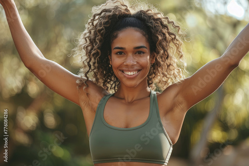 A fitness model in an olive green athleisure outfit, showcasing her toned arm muscles with arms raised against the natural backdrop of lush trees and sunlight