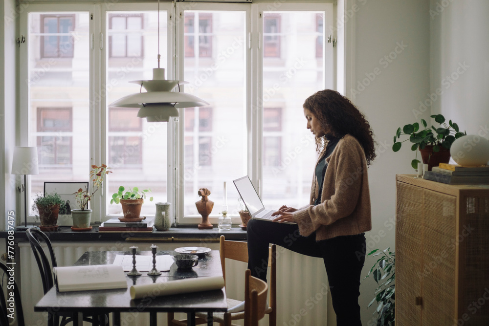 Side view of female construction engineer using laptop while standing with one leg on chair near table at home Side view of female construction engineer using laptop while standing with one leg on chair near table at home