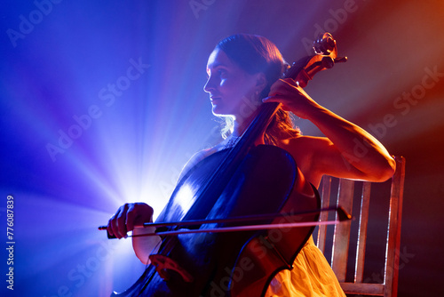 Cellist immersed in music, with striking halo of stage lights accentuating her silhouette on stage. Classic music in modern life. Concept of hobby and work, music festivals, concerts, symphony show.