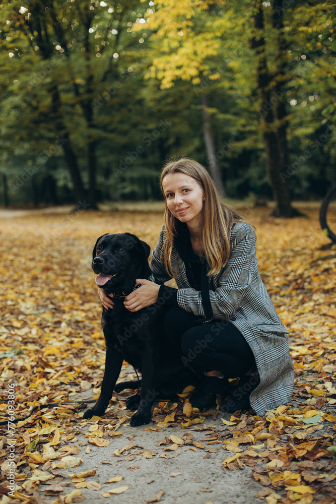 woman with a black labrador dog in the autumn park. walking with a dog in the park. hug and love the dog. a dog is a best friend. golden autumn and fallen leaves in the park