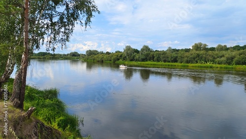 A motorized plastic boat floats on the river and a fisherman catches predatory fish by trolling. Illuminated by the summer sun, birch branches lean over the water. Grass, trees and bushes grow