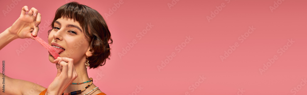 happy girl with nose piercing licking sweet and sour candy strip on pink background, horizontal - obrazy, fototapety, plakaty 